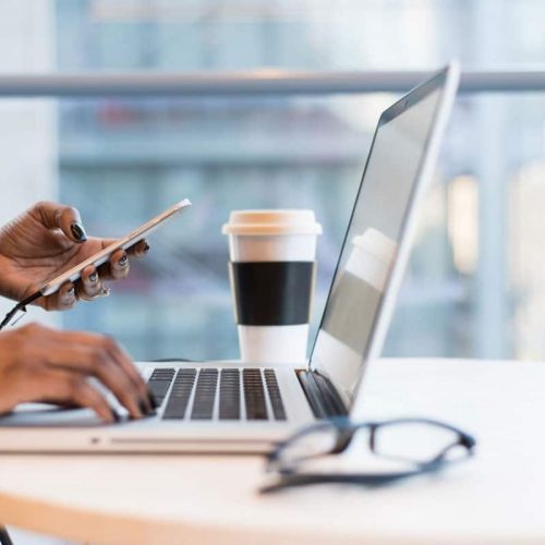 Two hands at a desk holding a mobile phone and working on a laptop.