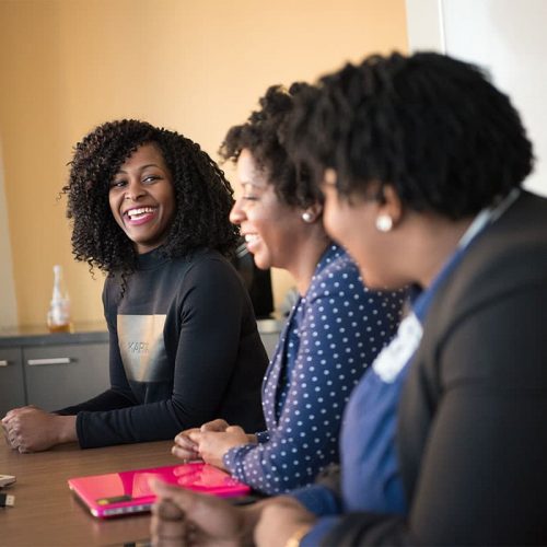 Three black women sitting in a row next to each other at a table, with one of them in focus and smiling at the others.