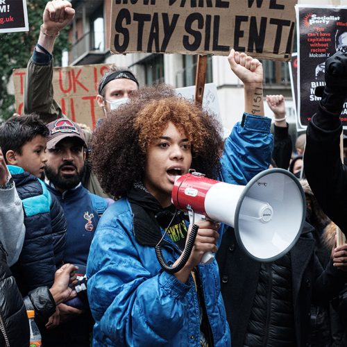 A young black woman wearing a blue jacked and holding a megaphone with her right fist in the air, standing with a group of anti-racism protestors.
